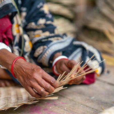 This image captures the skilled hands of a woman in Bangladesh engaged in traditional craftsmanship. Her fingers, adorned with vibrant red and white bangles, meticulously weave thin strips of bamboo or similar natural fibers. She is creating what appears to be a mat or a basket, a common and essential craft in many parts of the country. The focus on her hands and the intricate work highlights the generational knowledge and dexterity involved in this form of art. The backdrop of her traditional attire further emphasizes the cultural significance of this age-old practice.
