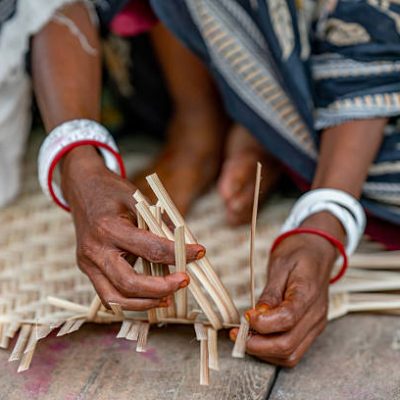 This image captures the skilled hands of a woman in Bangladesh engaged in traditional craftsmanship. Her fingers, adorned with vibrant red and white bangles, meticulously weave thin strips of bamboo or similar natural fibers. She is creating what appears to be a mat or a basket, a common and essential craft in many parts of the country. The focus on her hands and the intricate work highlights the generational knowledge and dexterity involved in this form of art. The backdrop of her traditional attire further emphasizes the cultural significance of this age-old practice.